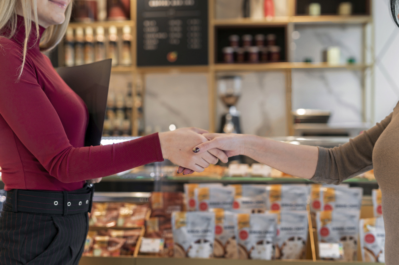 Deux femmes se serrent la main à l'intérieur d'un magasin moderne, avec des étagères de produits emballés et une machine à café en arrière-plan.