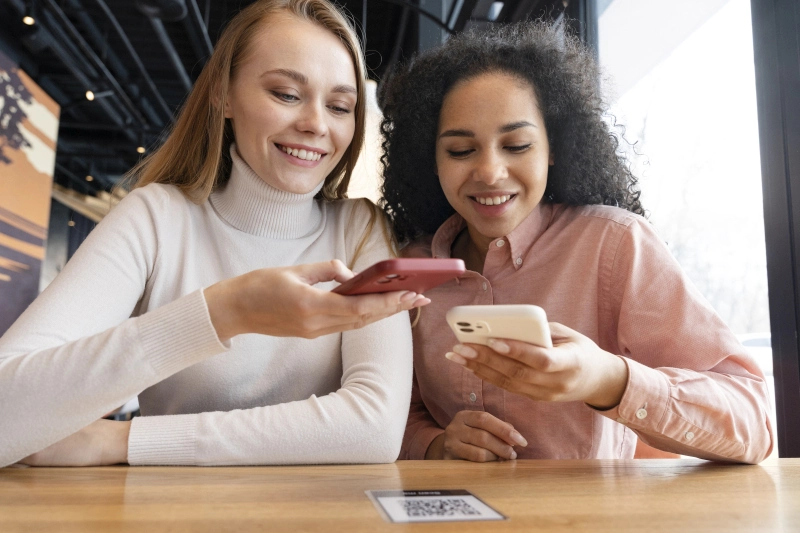 Zwei lächelnde Frauen, die an einem Cafétisch sitzen und mit ihren Smartphones einen QR-Code scannen, mit dem sie wahrscheinlich auf ein Kundenbindungsprogramm oder eine digitale Speisekarte zugreifen.