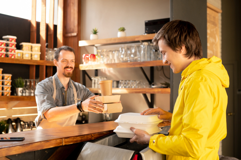Un barista souriant remet à un client vêtu d'une veste jaune, au comptoir d'un café confortable, une commande à emporter comprenant des récipients et du café.