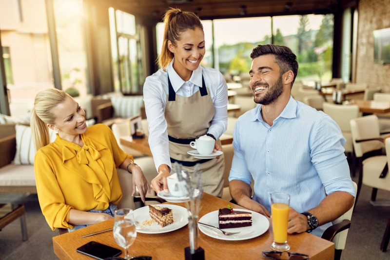 Une serveuse souriante sert du café à un homme et une femme joyeux assis à une table de restaurant avec des tranches de gâteau et des boissons.