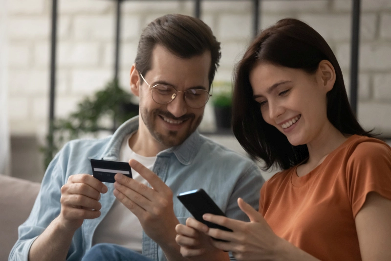 ein lächelndes Paar, das auf einer Couch sitzt, ein Smartphone und eine Kreditkarte in der Hand hält und gemeinsam ein Kundenbindungsprogramm durchsucht