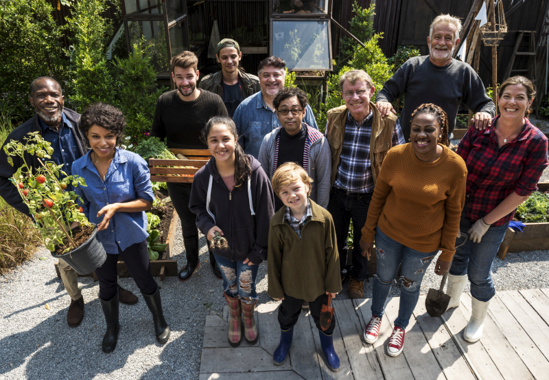Un groupe diversifié de personnes souriantes de tous âges se tient dans un jardin, certaines tenant des plantes et des outils de jardinage.